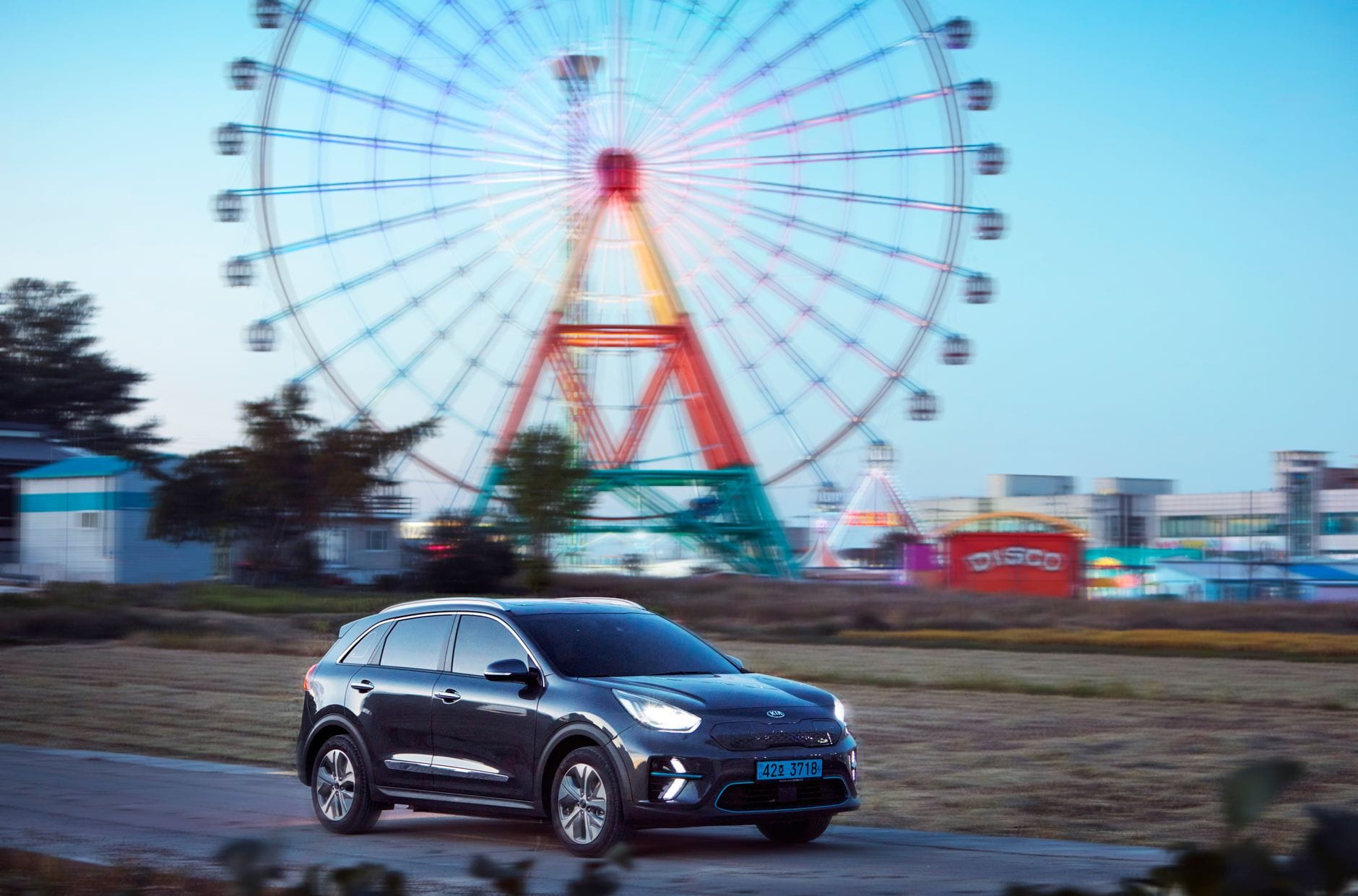 Elektryczny SUV nocą mija diabelski młyn w wesołym miasteczku Elektryczny SUV nocą mija diabelski młyn w wesołym miasteczku